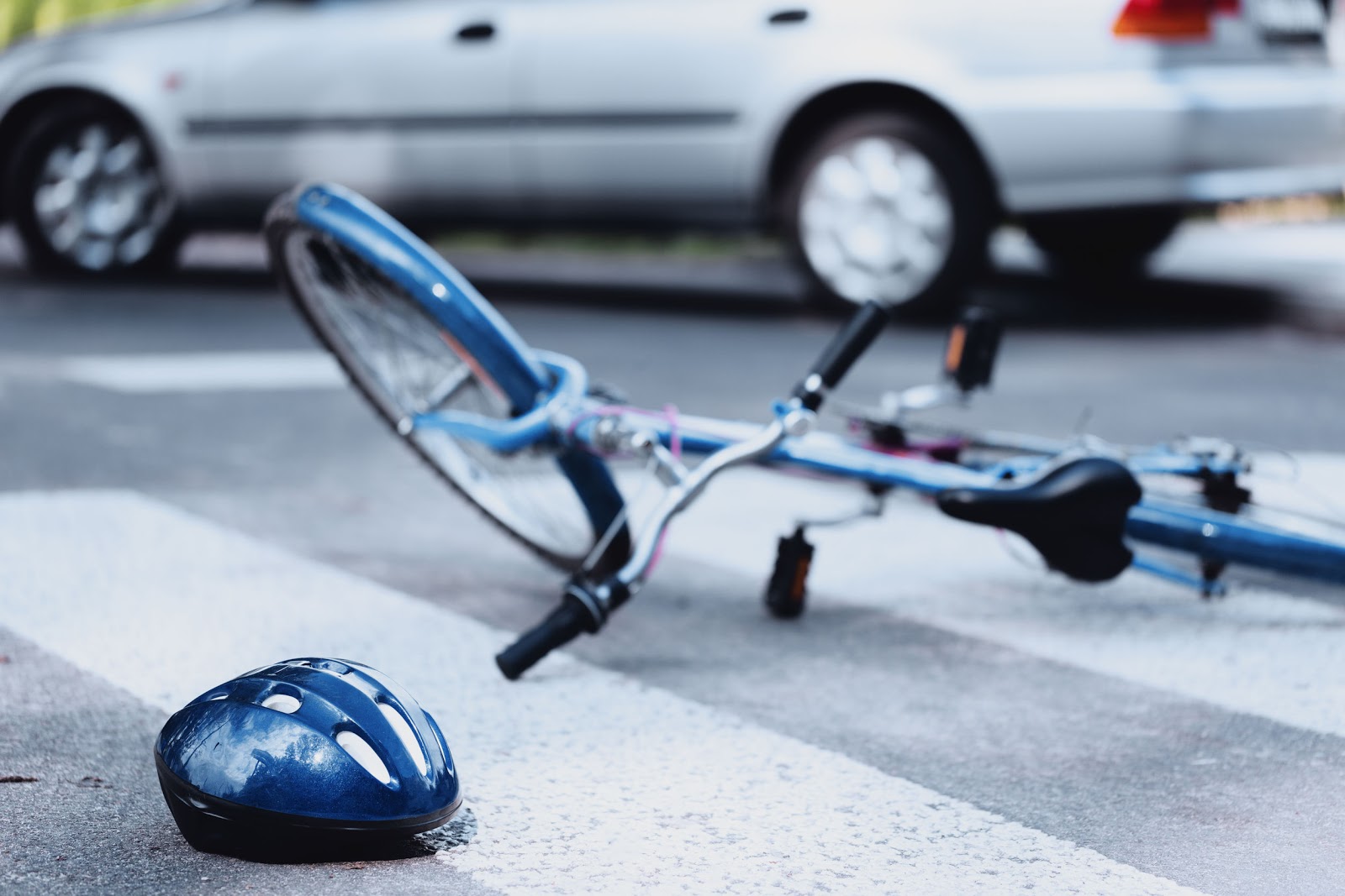 Scene of a bicycle accident at a crosswalk, with a blue helmet and bicycle lying on the ground and a car in the background, suggesting potential injuries and the need for legal assistance.