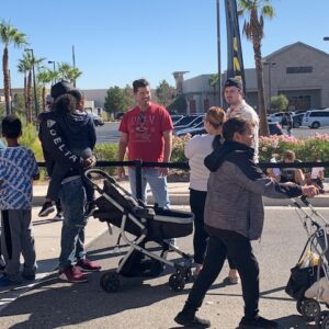 A diverse group of people are gathered outside on a sunny day in Las Vegas, some with strollers, depicting a community event where personal injury can occur.