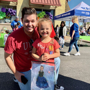 A Las Vegas personal injury attorney smiles with a young girl holding a costume at a community event with a tent that says "PAULPOWELL.COM (702) 728-5500," balloons, tables and other people in the background.