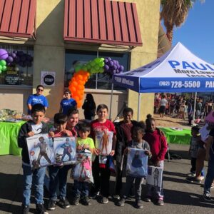A group of children stand in front of a tent for a Las Vegas Personal Injury Law Firm, holding toys and gifts at a community event.