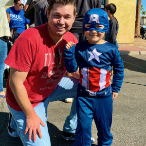A man smiles next to a child dressed as Captain America at a community event, showcasing support for families in need of personal injury legal services in Las Vegas.