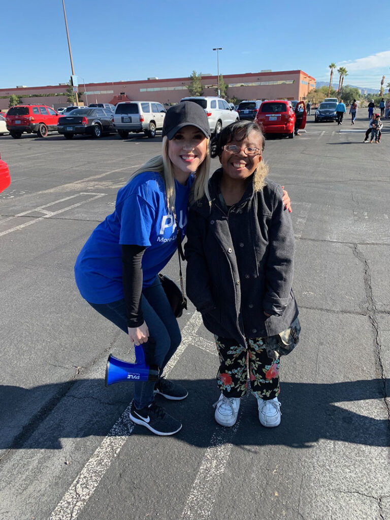 A woman in a blue shirt with a logo embraces a woman in a parking lot, representing community outreach and support from a personal injury law firm in Las Vegas.