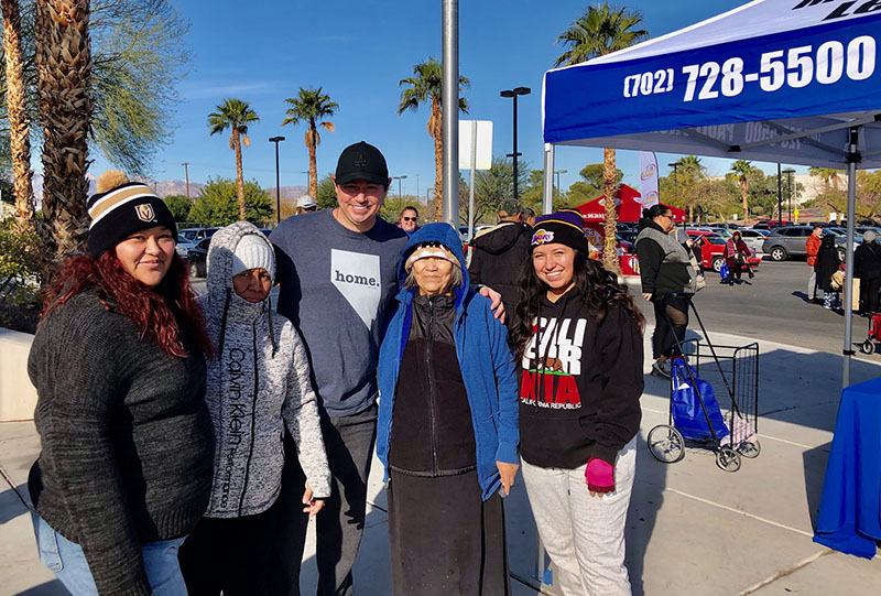 Community outreach event by a Las Vegas personal injury law firm, featuring a lawyer posing with local community members under a tent with the firm's phone number visible, set against a sunny Las Vegas backdrop with palm trees.
