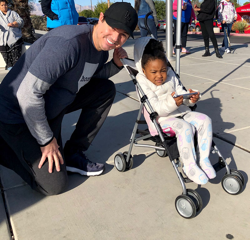 Las Vegas attorney kneels beside a young girl in a stroller, demonstrating community support and emphasizing care beyond personal injury claims.