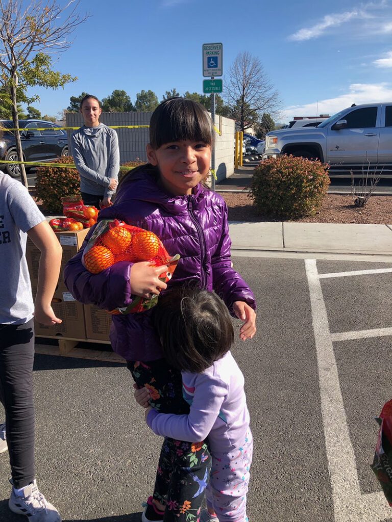 A smiling girl holds oranges while a younger girl hugs her, with a reserved parking sign in the background, potentially symbolizing accessible support after a personal injury in Las Vegas.
