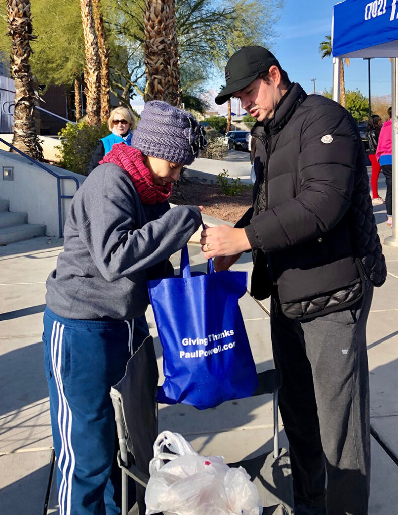 Man helping a woman with a blue tote bag labeled "Giving Thanks PaulPowell.com" at a community event, ideal for a Las Vegas personal injury law firm.