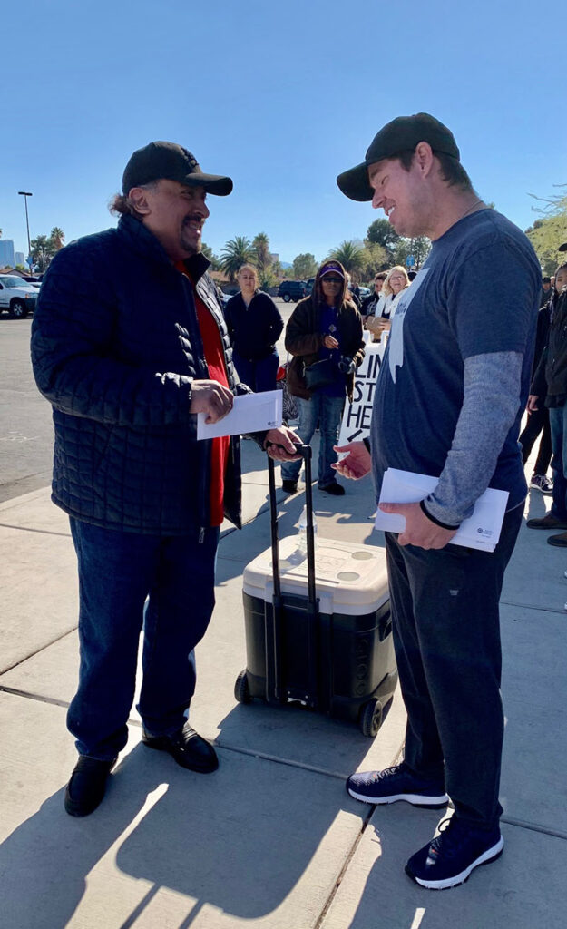 Two men exchanging envelopes in Las Vegas, likely compensation checks for personal injury claims, with a crowd of people in the background.