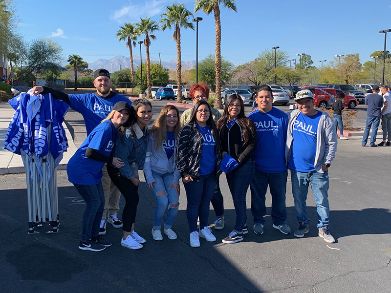 A group of the Paul Law Firm team in Las Vegas are gathered outside on a sunny day, wearing matching blue shirts with the firm's logo, ready for a community event.
