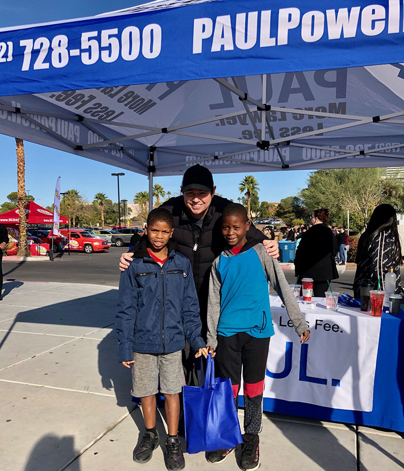 Image of Paul Powell, a personal injury attorney in Las Vegas, posing with two young boys under a tent displaying his firm's name and contact information, likely at a community event.