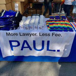 Promotional table with tote bags, mugs, rainbow-themed items, and the slogan "More Lawyer. Less Fee. PAUL." for a Las Vegas personal injury law firm.