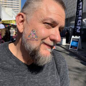 Man with a "Paul Powell Pride" unicorn tattoo on his cheek smiles in a sunny outdoor setting, possibly at a Las Vegas event, showcasing approachability and a connection to the community for a personal injury law firm.