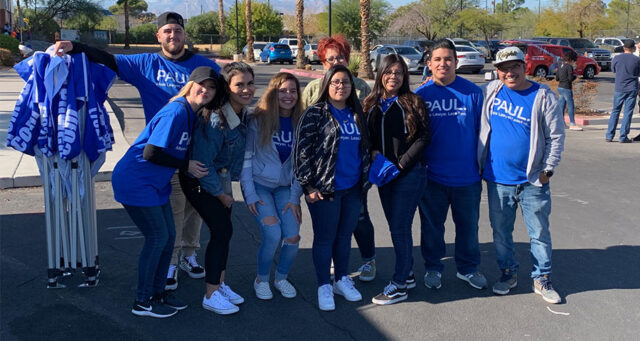 A group of staff members from a Personal Injury Law Firm in Las Vegas pose outdoors wearing blue shirts with their law firm's logo, with cars and palm trees visible in the background, representing their active role in the community.