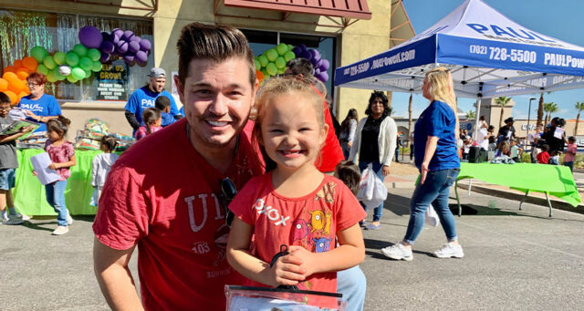 A man and a young girl smile at a community event hosted by a Las Vegas personal injury law firm, featuring books and a balloon arch.
