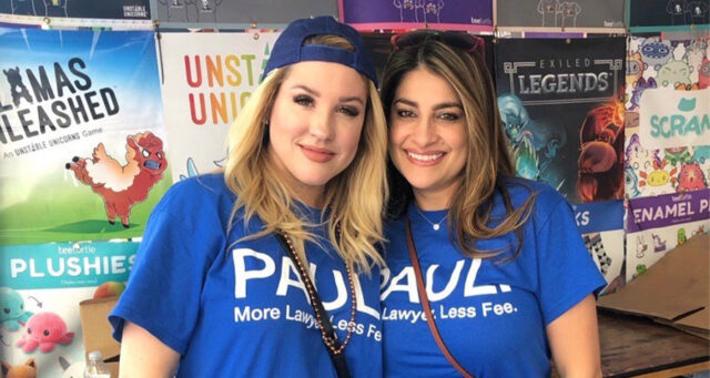 Two smiling women wearing matching blue "Paul: More Lawyer Less Fee" t-shirts at a community event for a Las Vegas personal injury law firm.