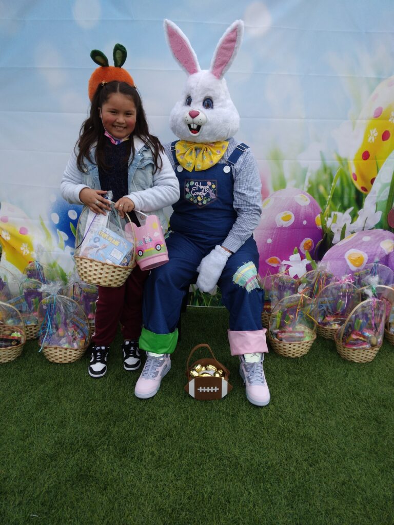 A young girl with a carrot headband smiles next to the Easter Bunny, surrounded by Easter baskets on a grass-like surface, with a painted Easter background, promoting family safety and personal injury services in Las Vegas.