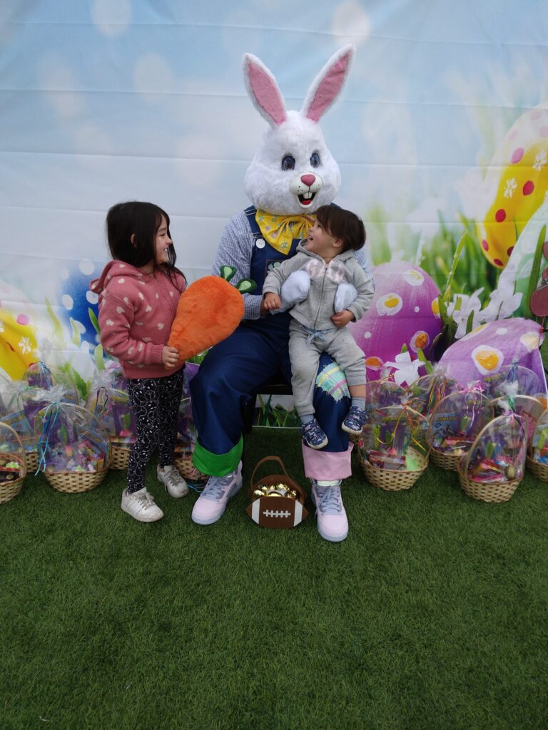The Easter Bunny poses with two children holding a carrot and a plush tail in front of Easter baskets at a community event hosted by a Las Vegas personal injury law firm, emphasizing family and community support.