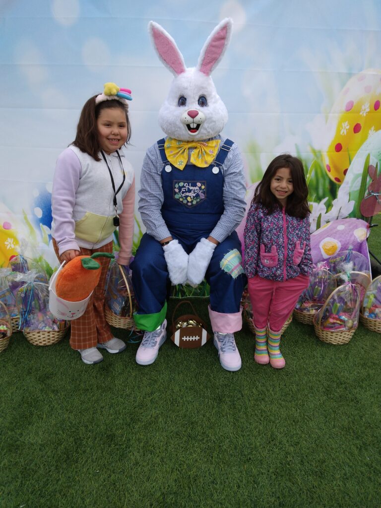 Two smiling young girls pose with the Easter Bunny, surrounded by Easter baskets on green grass, against a backdrop of decorated eggs; image for a Las Vegas personal injury law firm promoting community involvement and family safety.