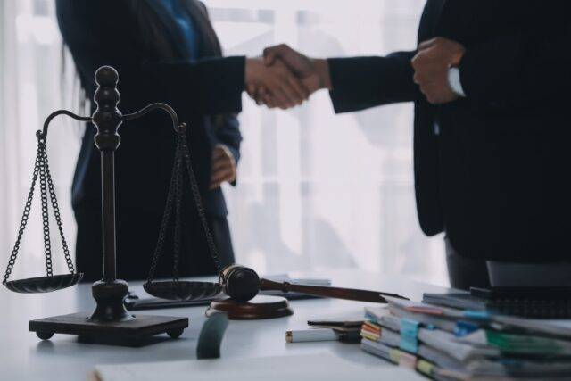 Close-up image of attorneys shaking hands after agreeing to represent a personal injury client, with scales of justice and a gavel in the foreground.