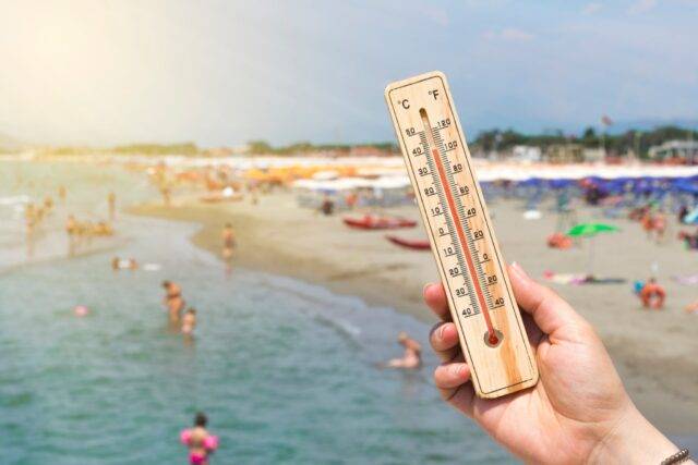 Hand holding a thermometer displaying high temperature with a crowded beach scene in the background, symbolizing the risk of heatstroke and the need for personal injury legal assistance in Las Vegas.