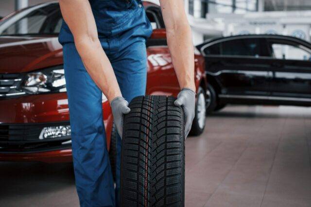 A mechanic in blue overalls and gloves holds a tire in a car repair shop, symbolizing the potential for tire defects or improper maintenance leading to car accidents and personal injury claims in Las Vegas.