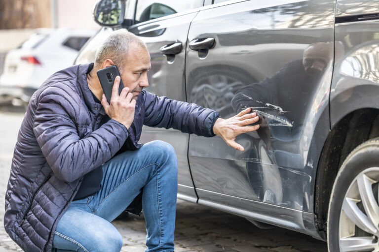 A man kneels next to a gray car, examining damage from a car accident and talking on his cell phone, suggesting a call to a Las Vegas personal injury law firm for assistance.