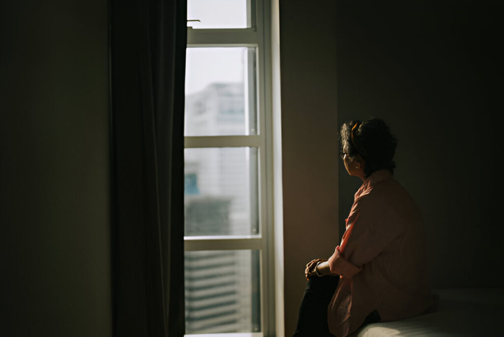 Lonely asian chinese senior woman standing in the dark behind the window looking