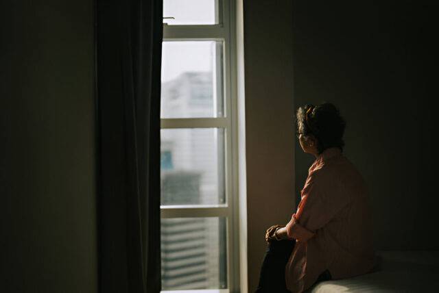 Lonely asian chinese senior woman standing in the dark behind the window looking